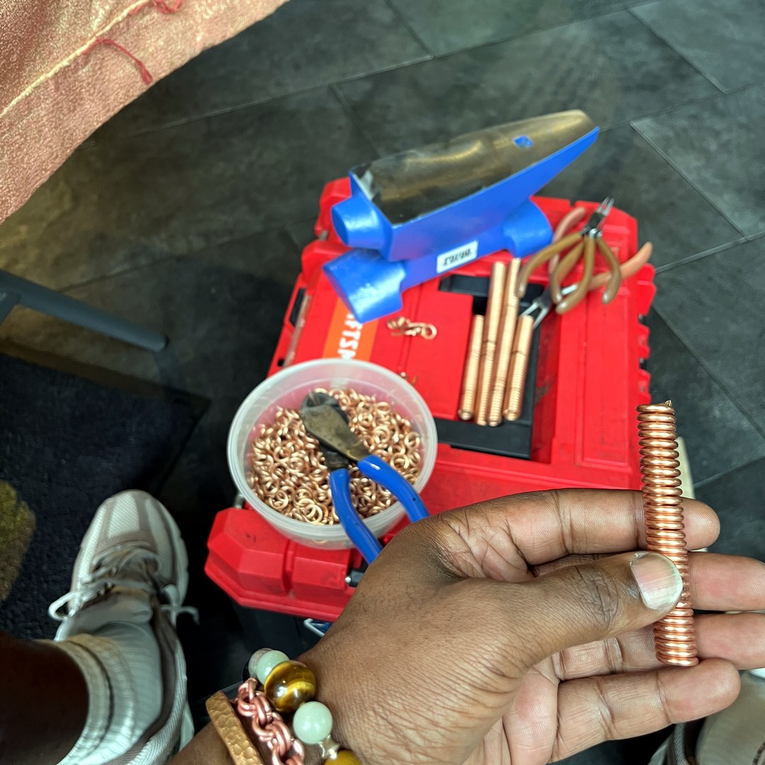 Baker's workshop floor — anvil, hammer, jump rings, copper wire stock, finished bracelets on his wrist