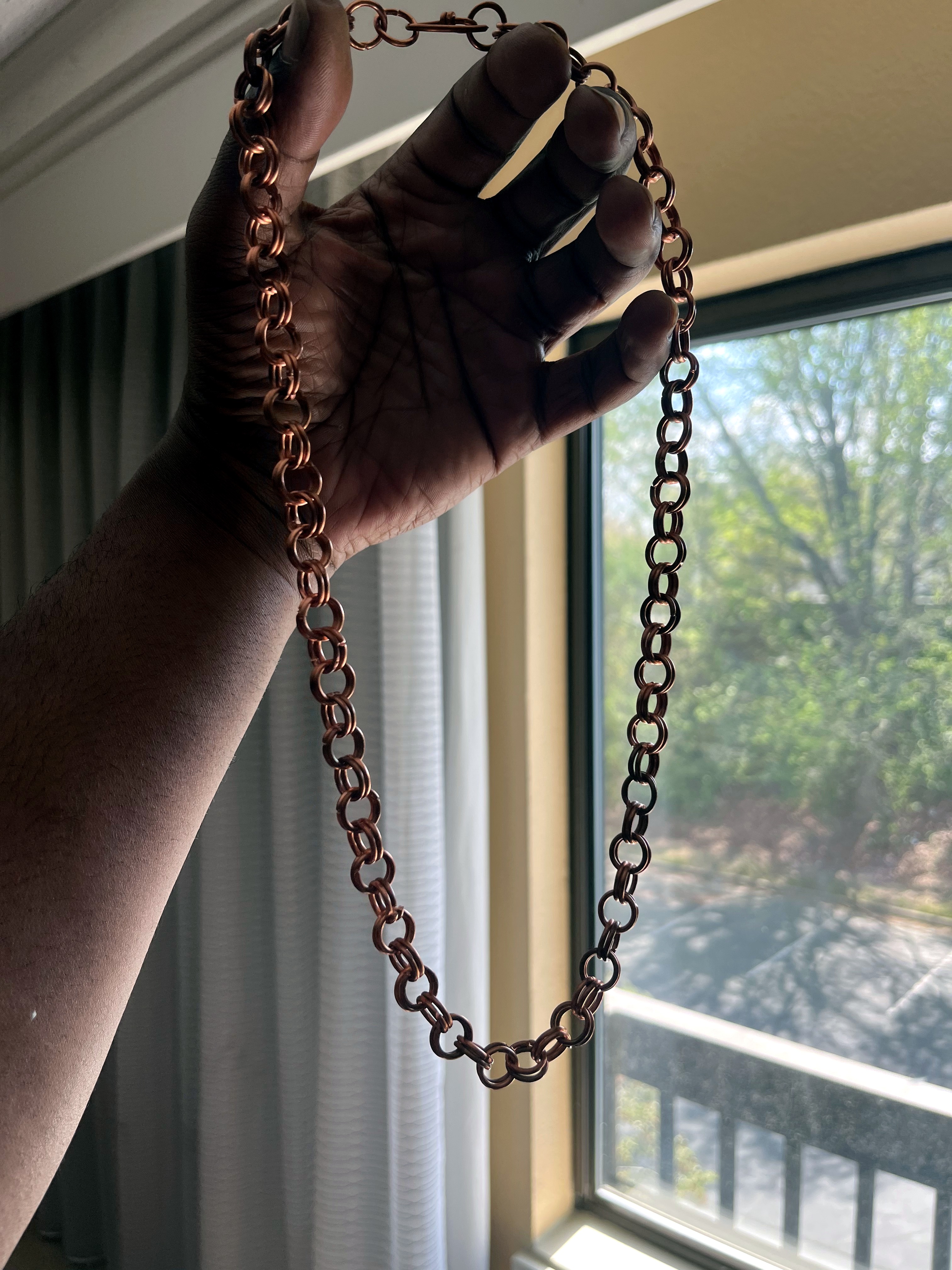 Long copper Byzantine chain held in hand, sunlight from window catching the metal