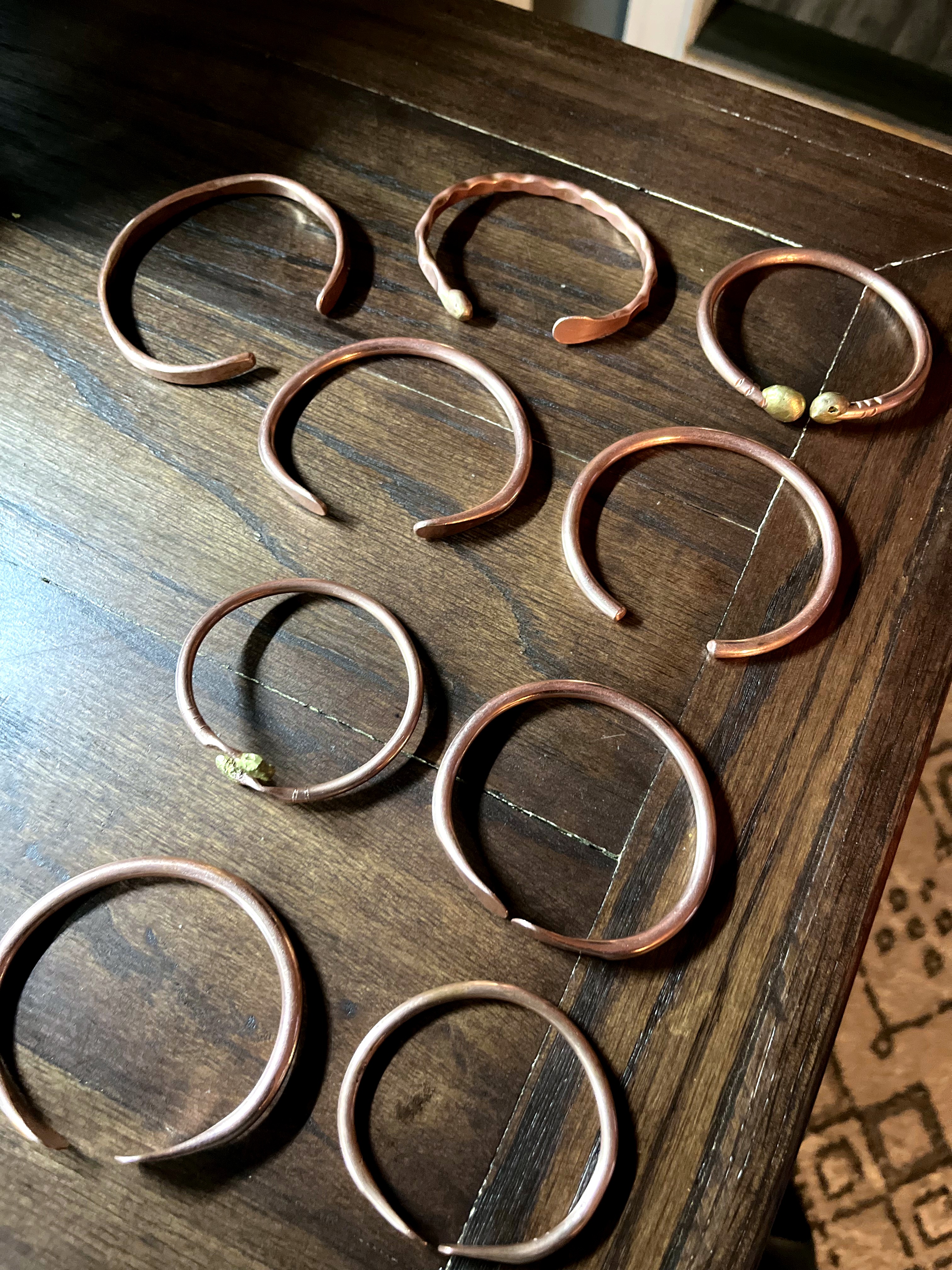 Nine hand-forged copper cuff bracelets arranged in a 3x3 grid on dark wood